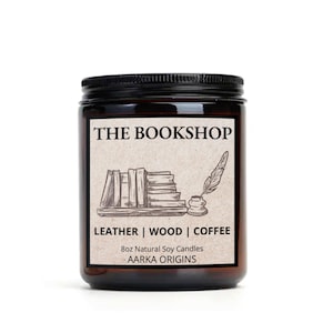 May include: A brown glass jar candle with a black lid. The label reads "THE BOOKSHOP" above a sketch of books, a quill, and inkwell. Below, it says "LEATHER | WOOD | COFFEE" and "8oz Natural Soy Candles AARKA ORIGINS."