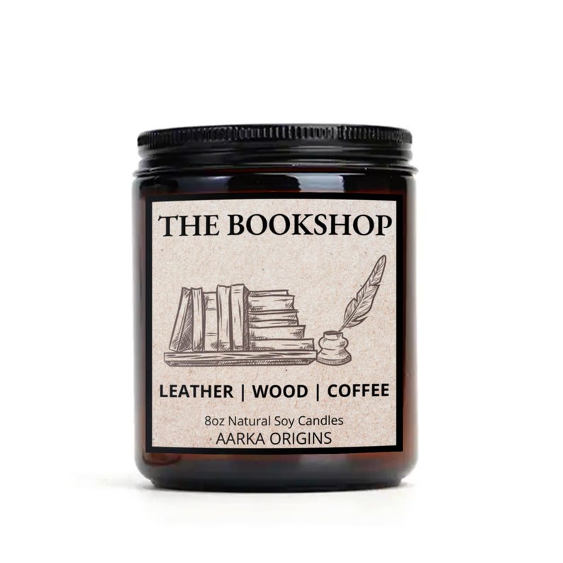 May include: A brown glass jar candle with a black lid. The label reads "THE BOOKSHOP" above a sketch of books, a quill, and inkwell. Below, it says "LEATHER | WOOD | COFFEE" and "8oz Natural Soy Candles AARKA ORIGINS."