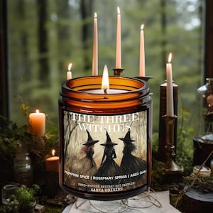 May include: A lit candle in a brown glass jar with a label that reads "THE THREE WITCHES." The label features an illustration of three witches in a forest. The candle is surrounded by other lit candles and a natural background.