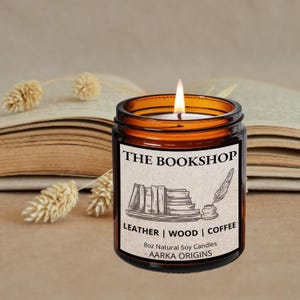 May include: A lit, amber-colored candle in a glass jar labeled "THE BOOKSHOP." The label features a sketch of books and a quill. The candle is 8oz and scented with leather, wood, and coffee. The candle sits in front of an open book.