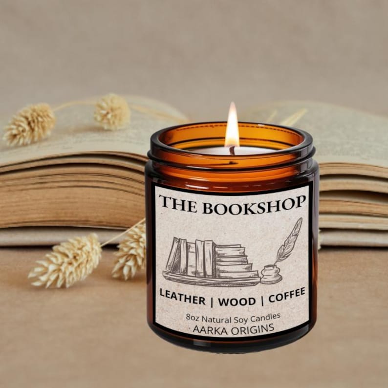 May include: A lit, amber-colored candle in a glass jar labeled "THE BOOKSHOP." The label features a sketch of books and a quill. The candle is 8oz and scented with leather, wood, and coffee. The candle sits in front of an open book.
