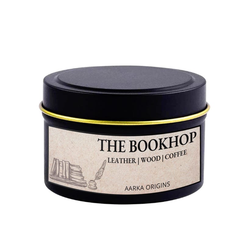 May include: A black round tin with a gold rim and a label that reads "THE BOOKHOP" with the scent notes "LEATHER | WOOD | COFFEE". The label also features a stack of books and a quill illustration. The brand name "AARKA ORIGINS" is at the bottom.