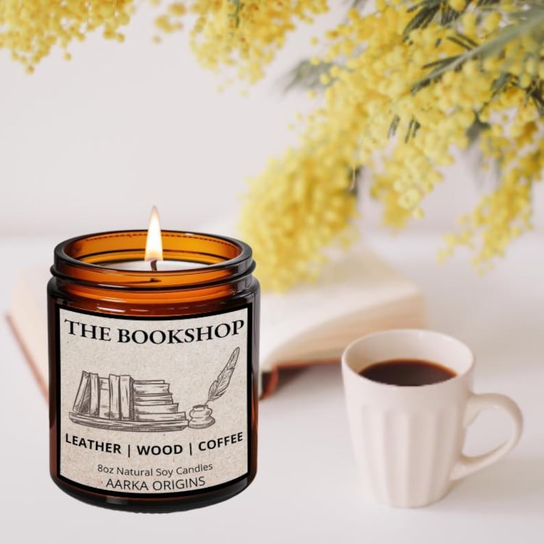 May include: Lit 8oz natural soy candle in a brown glass jar with a label that reads "THE BOOKSHOP" with illustrations of books and a quill. The candle's scent is Leather, Wood, and Coffee. A white mug of coffee sits nearby.