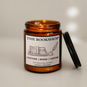 May include: An amber-colored glass jar candle with a black lid. The label reads "THE BOOKSHOP" with illustrations of books and a quill. The candle is labeled "LEATHER | WOOD | COFFEE" and is 8oz.