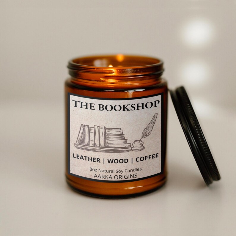 May include: An amber-colored glass jar candle with a black lid. The label reads "THE BOOKSHOP" with illustrations of books and a quill. The candle is labeled "LEATHER | WOOD | COFFEE" and is 8oz.