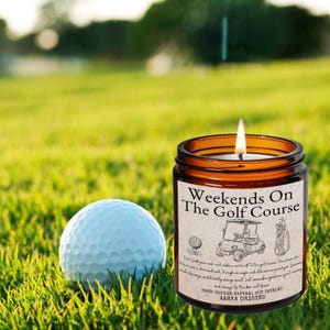May include: A lit candle in a brown glass jar with the text "Weekends On The Golf Course" and golf-themed illustrations. A white golf ball sits on the green grass next to the candle. The candle is a hand-poured natural soy candle.