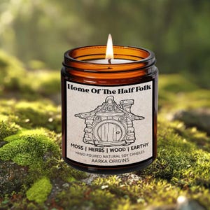 May include: A lit, amber-colored glass jar candle with a label that reads "Home Of The Half Folk." The label features a line drawing of a hobbit hole. The candle's scent is described as moss, herbs, wood, and earthy. The candle is hand-poured natural soy.