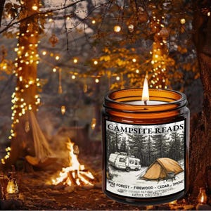 May include: A lit candle in a brown glass jar with the text "CAMPSITE READS" on the label. The label features a black and white illustration of a camper and tent in a forest. The candle is labeled "PINE FOREST - FIREWOOD - CEDAR - SMOKE".