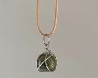 May include: A green stone pendant encased in a silver wire cage, hanging from a beige cord necklace.