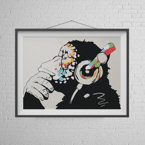 May include: Framed art print featuring a stylized chimpanzee wearing headphones. The chimpanzee is rendered in black and white, with colorful accents on the face and headphones. The artwork is displayed in a black frame against a white brick wall.