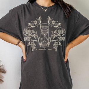 May include: A dark gray oversized t-shirt with a graphic print of an hourglass, butterflies, and roses. The text "memento mori" is printed below the hourglass.
