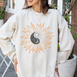 May include: A cream colored sweatshirt with a graphic of a yin yang symbol inside of a sunflower.