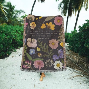 May include: A woven tapestry throw blanket with a dark background and a floral design featuring pressed flowers. The blanket has a quote from Isaiah 40:8, "The grass withers, the flower fades, but the word of our God will stand forever."