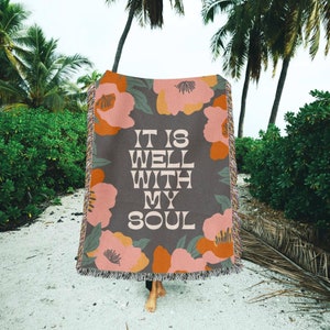 May include: A woven throw blanket with a floral pattern in shades of pink, orange, and green. The blanket has the text "It is well with my soul" in a retro font.