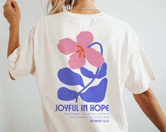 May include: Cream-coloured t-shirt with a pink and blue floral design. The shirt displays the text "JOYFUL IN HOPE" with further text below, referencing Romans 12:12. The design is on the back of the shirt.