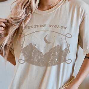 Może przedstawiać: Jasnobeżowy t-shirt z nadrukiem graficznym przedstawiającym pasmo g&oacute;rskie, sierp księżyca i gwiazdy. Tekst "WESTERN NIGHTS" jest nadrukowany nad grafiką.