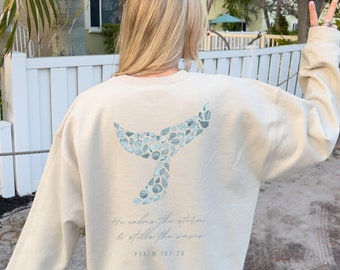 May include: Cream-colored sweatshirt featuring a whale tail design composed of blue and white seashells. The text "He calms the storm & stills the waves" and "Psalm 107:29" are printed below the whale tail. The sweatshirt is a relaxed fit.