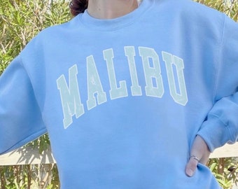 Op de afbeelding: Een lichtblauw sweatshirt met het woord "MALIBU" in lichtgroen gedrukt op de voorkant. Het sweatshirt wordt gedragen door een persoon die buiten staat in de buurt van een auto.