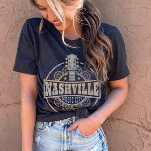 Puede incluir: Una camiseta gris oscuro con un gráfico de estilo vintage de una guitarra y el texto "Nashville Music City".