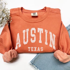 May include: A burnt orange sweatshirt with the text "AUSTIN TEXAS" in white block letters.