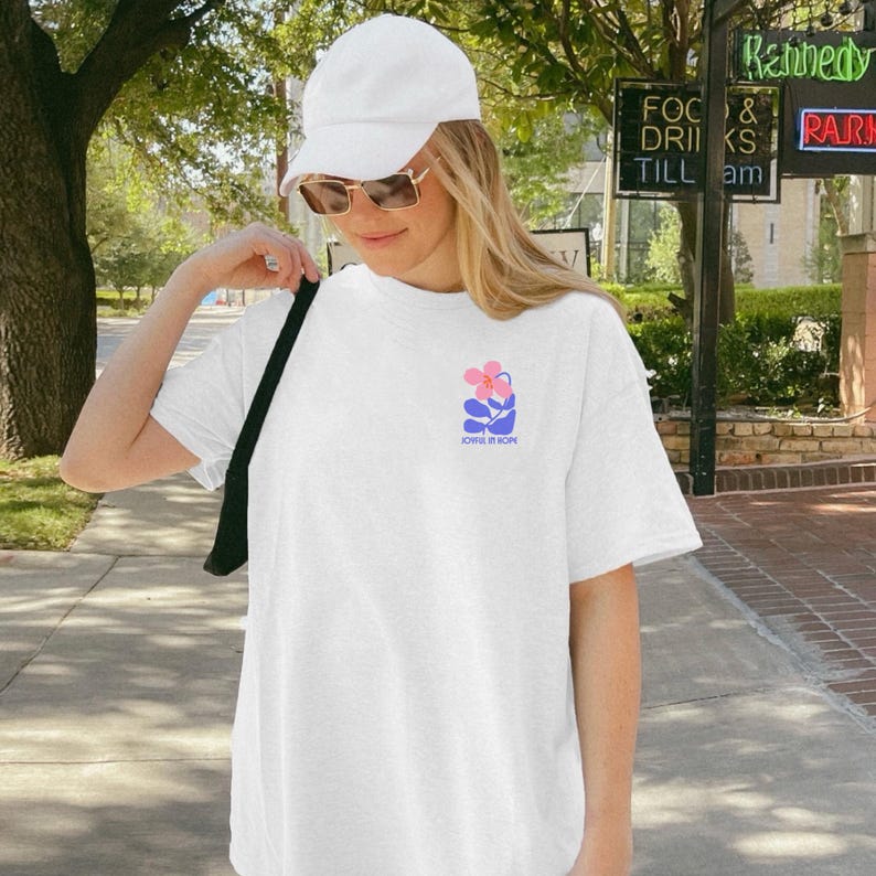 Op de afbeelding: Wit T-shirt met een roze en blauwe bloemenprint en de tekst "JOIN IN HOPE". Het shirt wordt gedragen met een witte pet en zonnebril. De foto is buiten gemaakt.