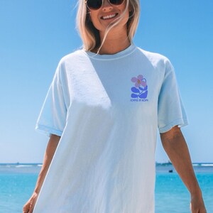 Op de afbeelding: Lichtblauw T-shirt met een bloemenontwerp in paars en blauw. De tekst "JOYFUL IN HOPE" is onder de afbeelding gedrukt. Het shirt is gemaakt van een zacht materiaal en heeft korte mouwen.