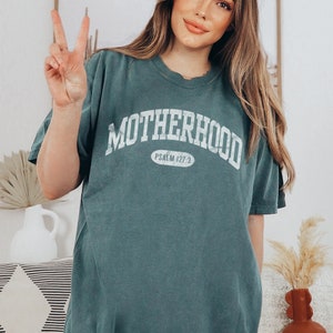 May include: A teal blue t-shirt with the word "MOTHERHOOD" in white text in an arched font. The text "PSALM 127:3" is in a smaller white font inside a white oval.