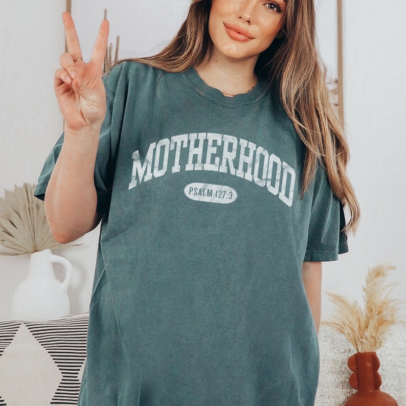May include: A teal blue t-shirt with the word "MOTHERHOOD" in white text in an arched font. The text "PSALM 127:3" is in a smaller white font inside a white oval.