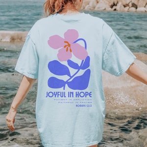 Op de afbeelding: Lichtblauw T-shirt met een bloemenontwerp in roze en blauw. De tekst "JOYFUL IN HOPE" staat eronder, samen met extra tekst uit Romeinen 12:12. Het shirt heeft een casual, oversized stijl.