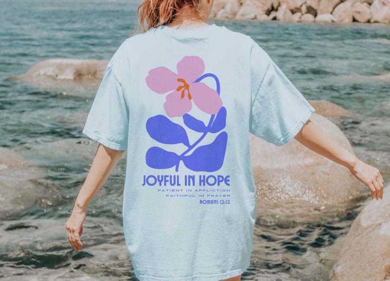 Op de afbeelding: Lichtblauw T-shirt met een bloemenontwerp in roze en blauw. De tekst "JOYFUL IN HOPE" staat eronder, samen met extra tekst uit Romeinen 12:12. Het shirt heeft een casual, oversized stijl.