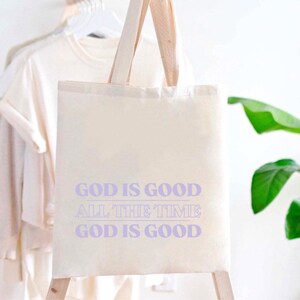 May include: A white canvas tote bag with purple text that reads "God is good all the time God is good".