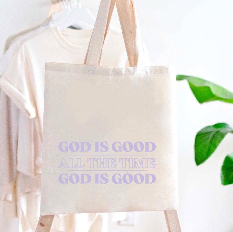 May include: A white canvas tote bag with purple text that reads "God is good all the time God is good".