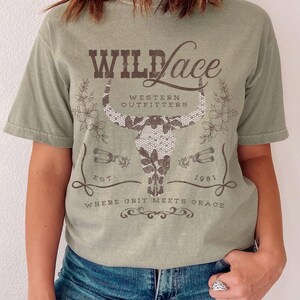 K&ouml;nnte beinhalten: Ein gr&uuml;nes T-Shirt mit einer braunen Grafik eines Bullensch&auml;dels mit einer Spitzen&uuml;berlagerung. Der Text "Wild Lace" befindet sich &uuml;ber dem Sch&auml;del und "Western Outfitters" darunter. Der Text "Est. 1981" befindet sich rechts neben dem Sch&auml;del und "Where Grit Meets Grace" darunter.