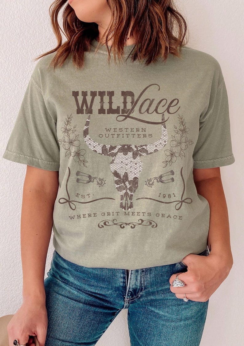 K&ouml;nnte beinhalten: Ein gr&uuml;nes T-Shirt mit einer braunen Grafik eines Bullensch&auml;dels mit einer Spitzen&uuml;berlagerung. Der Text "Wild Lace" befindet sich &uuml;ber dem Sch&auml;del und "Western Outfitters" darunter. Der Text "Est. 1981" befindet sich rechts neben dem Sch&auml;del und "Where Grit Meets Grace" darunter.