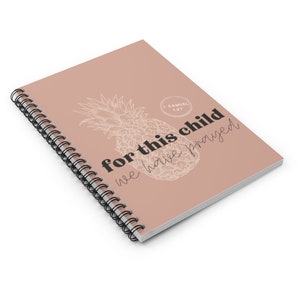 Puede incluir: Cuaderno con espiral y cubierta rosa rubor. La cubierta presenta un dise&ntilde;o de pi&ntilde;a esbozado y el texto "for this child we have prayed" en negro y fuente de escritura. Tambi&eacute;n se incluye el vers&iacute;culo b&iacute;blico "1 Samuel 1:27".