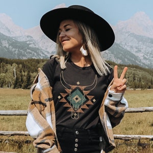 May include: A woman wearing a black t-shirt with a geometric design of a bull skull in green, brown, and gold. She is wearing a brown and white plaid shirt over the t-shirt and a black wide-brimmed hat. She is standing in a field with mountains in the background.