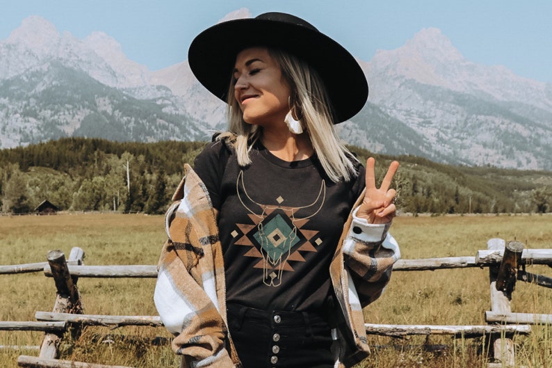 May include: A woman wearing a black t-shirt with a geometric design of a bull skull in green, brown, and gold. She is wearing a brown and white plaid shirt over the t-shirt and a black wide-brimmed hat. She is standing in a field with mountains in the background.