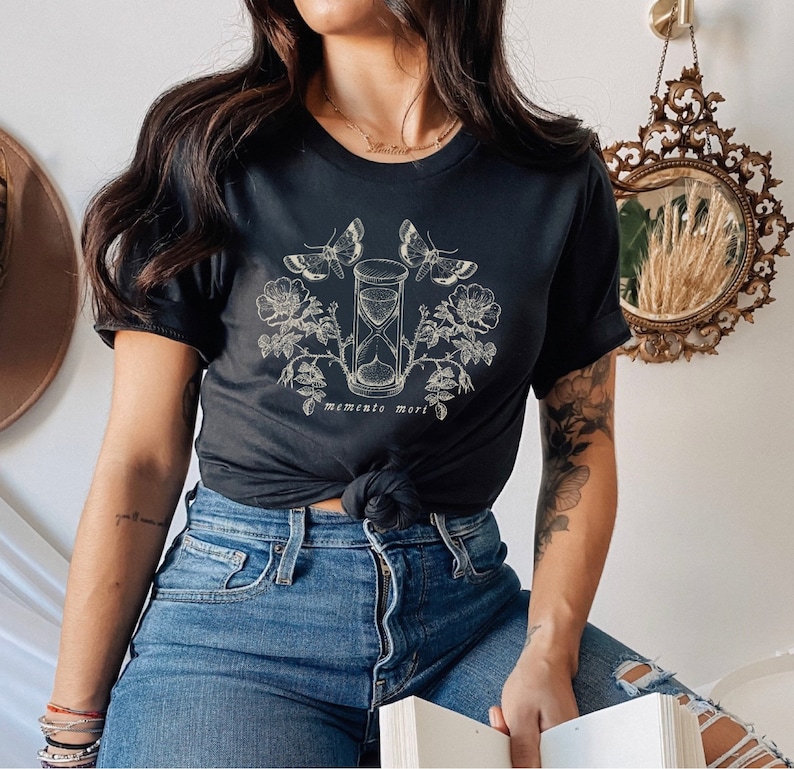 May include: Black t-shirt with a graphic of an hourglass, butterflies, and flowers. The text "memento mori" is printed below the hourglass.