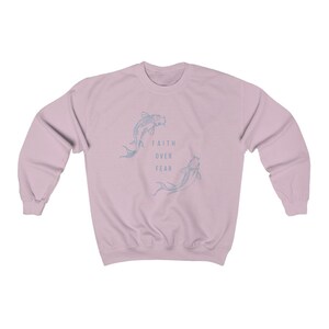 Puede incluir: Una sudadera de color rosa claro con un gr&aacute;fico de dos peces nadando en c&iacute;rculo. El texto "FAITH OVER FEAR" est&aacute; impreso en el centro del c&iacute;rculo.