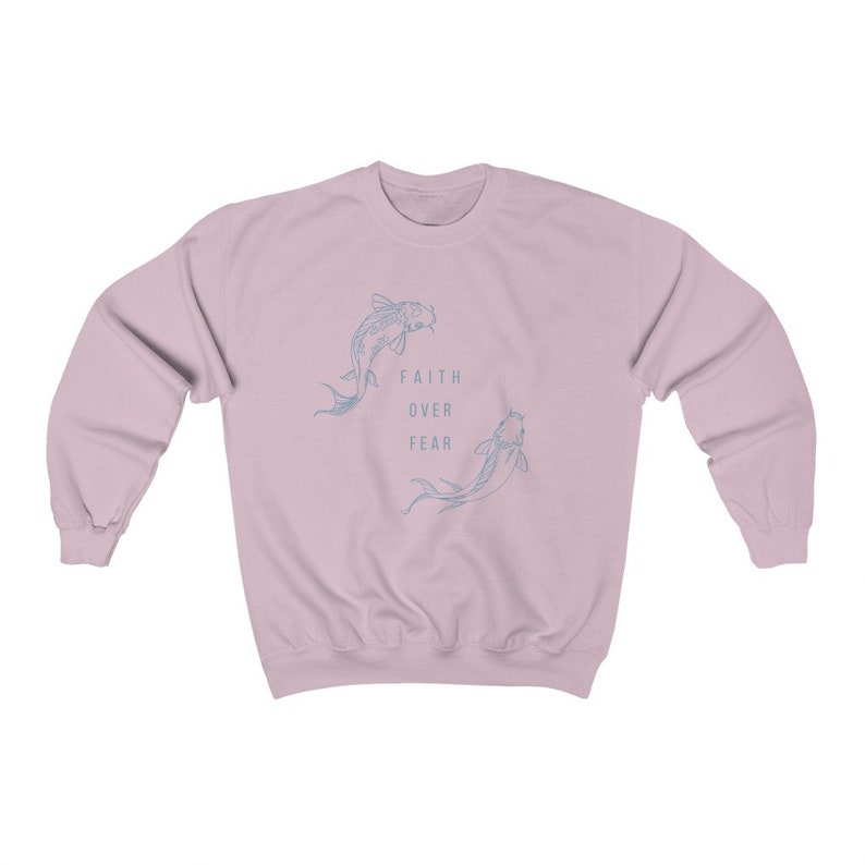 Puede incluir: Una sudadera de color rosa claro con un gr&aacute;fico de dos peces nadando en c&iacute;rculo. El texto "FAITH OVER FEAR" est&aacute; impreso en el centro del c&iacute;rculo.