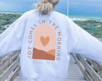 Könnte beinhalten: Weißer Pullover mit einem Grafikdruck eines rosafarbenen Bogens mit einem Herzen und Sonnenstrahlen. Der Text "Joy comes in the morning" ist vertikal auf der linken Seite des Bogens geschrieben und "Psalm 30:5" steht unter dem Bogen.