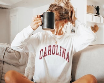 May include: A white long-sleeved sweatshirt with the word "CAROLINA" printed in maroon on the front. The sweatshirt is being worn by a person holding a black coffee mug.