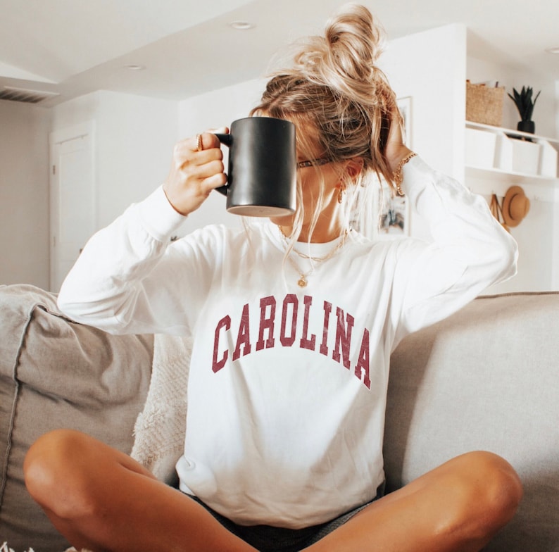 May include: A white long-sleeved sweatshirt with the word "CAROLINA" printed in maroon on the front. The sweatshirt is worn by a person holding a black coffee mug.