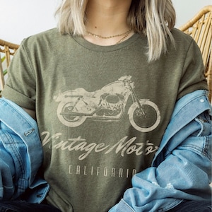 May include: A woman wearing a green t-shirt with a vintage motorcycle graphic and the text "Vintage Motors California". She is wearing a denim jacket and jeans.