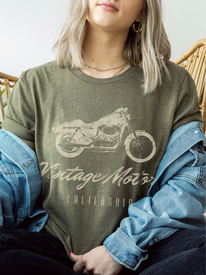 May include: A woman wearing a green t-shirt with a vintage motorcycle graphic and the text "Vintage Motors California". She is wearing a denim jacket and jeans.