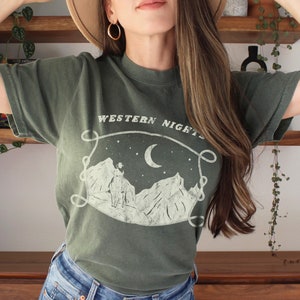 Może przedstawiać: Zielony T-shirt z białym nadrukiem przedstawiającym pasmo g&oacute;rskie, p&oacute;łksiężyc i gwiazdy. Na koszulce nadrukowany jest tekst "WESTERN NIGHTS".