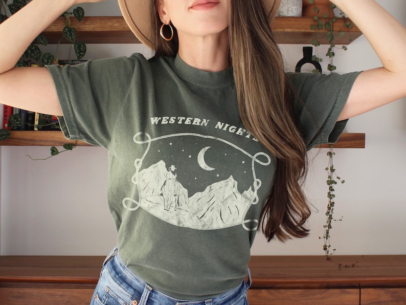 Może przedstawiać: Zielony T-shirt z białym nadrukiem przedstawiającym pasmo g&oacute;rskie, p&oacute;łksiężyc i gwiazdy. Na koszulce nadrukowany jest tekst "WESTERN NIGHTS".