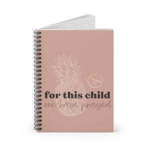 Puede incluir: Un cuaderno con espiral y tapa marr&oacute;n claro. Presenta un dise&ntilde;o de pi&ntilde;a esbozado y el texto "for this child we have prayed". El cuaderno tiene p&aacute;ginas rayadas y encuadernaci&oacute;n en espiral negra.
