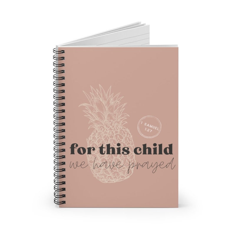 Puede incluir: Un cuaderno con espiral y tapa marr&oacute;n claro. Presenta un dise&ntilde;o de pi&ntilde;a esbozado y el texto "for this child we have prayed". El cuaderno tiene p&aacute;ginas rayadas y encuadernaci&oacute;n en espiral negra.