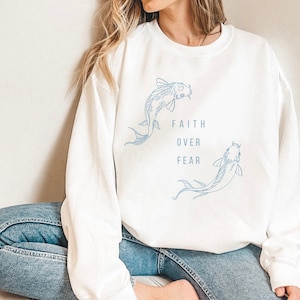 Puede incluir: Sudadera blanca con un contorno azul de dos peces koi y el texto "FAITH OVER FEAR".